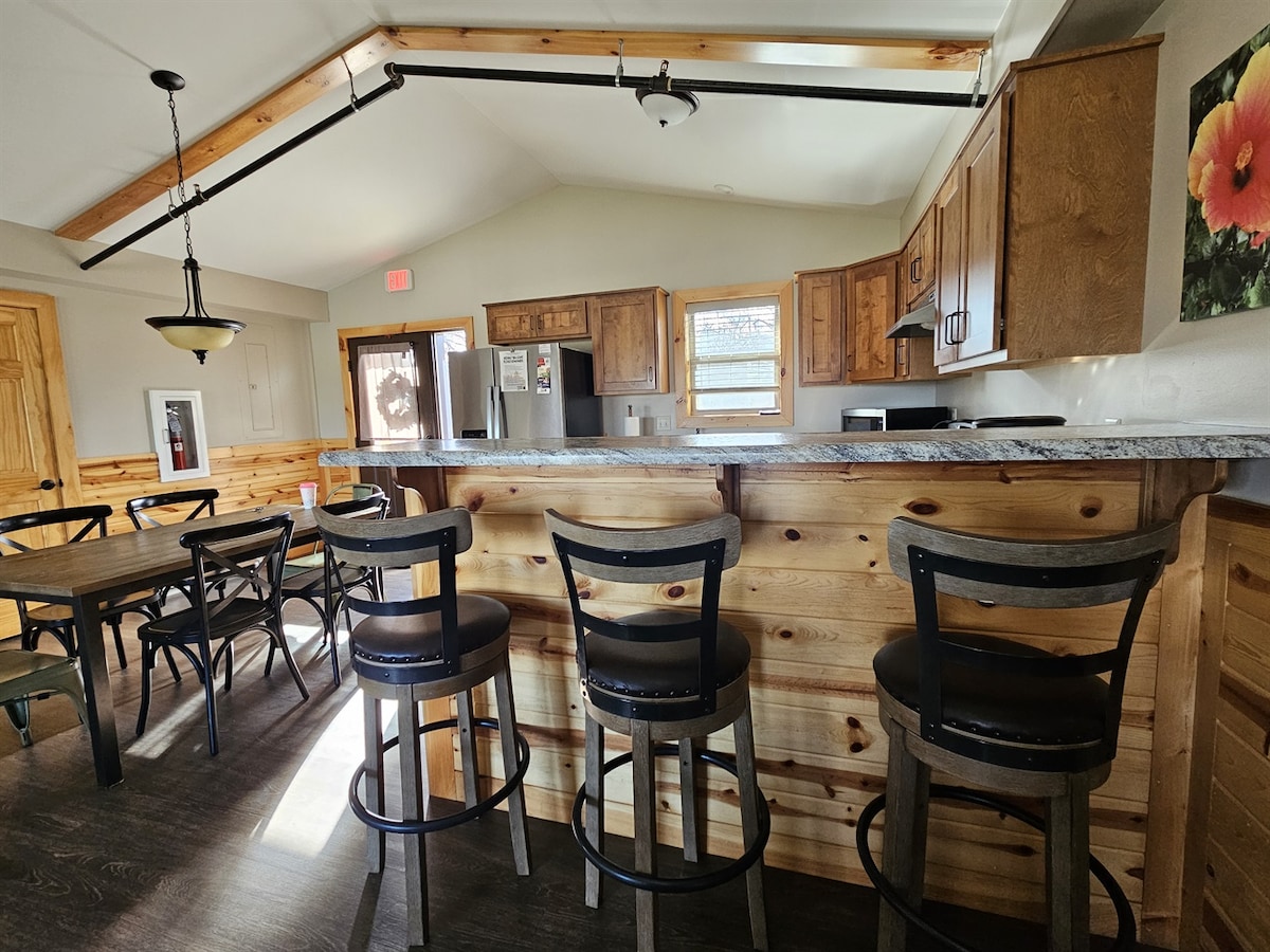 The kitchen area features wooden cabinetry and a central island with seating for four. A dining table for six is positioned nearby, creating a functional space for meals. Bright light streams through multiple windows, highlighting the room's open layout.