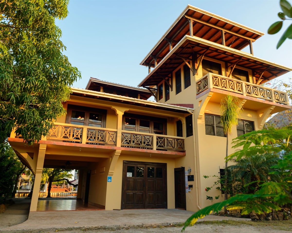 The exterior of the villa features a three-story structure with large, wooden balconies and a welcoming entrance. Lush greenery surrounds the property, enhancing its tropical feel. The building's architecture highlights open outdoor spaces, with ample windows allowing natural light to fill the interior.