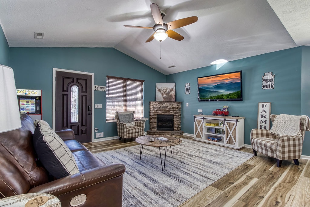 A spacious living area features comfortable seating arrangements, including a leather sofa and armchairs. A stone fireplace serves as a focal point beneath a mounted television displaying scenic mountain views. Light wood flooring complements the room's warm color palette, enhancing the cozy ambiance.
