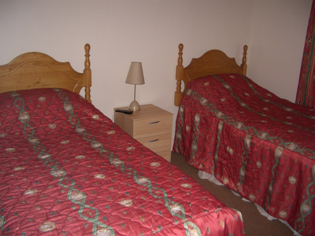 The room features two wooden beds with matching patterned bedspreads in shades of red. A small bedside table with a lamp is positioned between the beds, complementing the decor. Light fabric curtains frame a window, contributing to a bright ambiance.