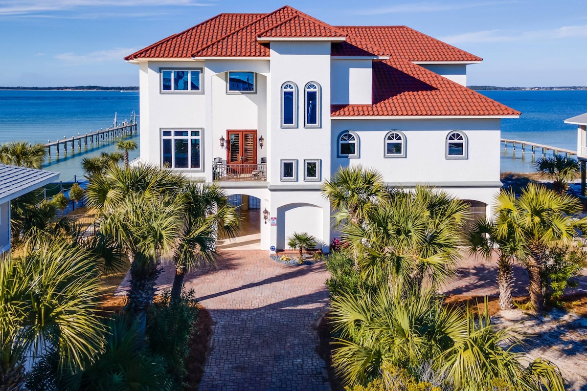 The exterior of the Mediterranean estate showcases a two-story structure with a red-tiled roof and large windows. Palm trees surround the property, leading to a private dock extending into the sparkling waters of the Santa Rosa Sound, creating a sense of tranquility and relaxation.