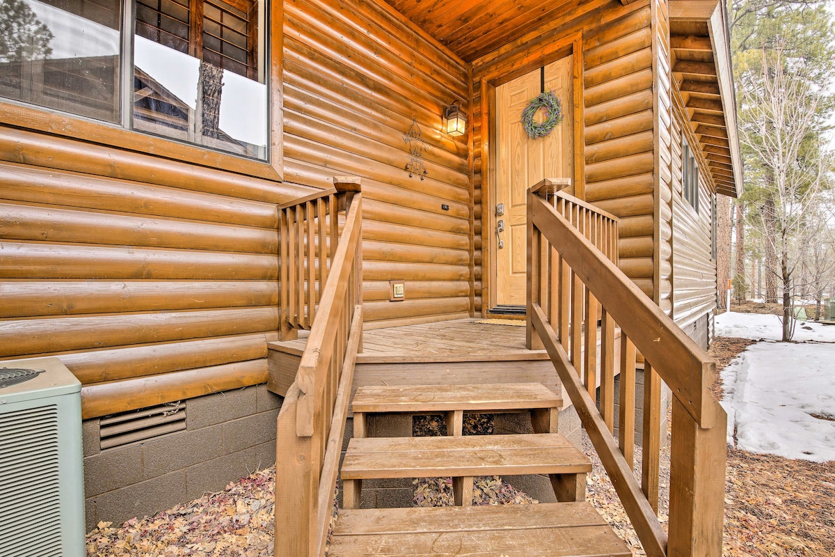 The entrance features a wooden deck with a set of five steps leading to a welcoming door. The exterior is constructed of log siding, complemented by a decorative wreath on the door. Surrounding the home, a winter scene highlights patches of snow and pine trees.