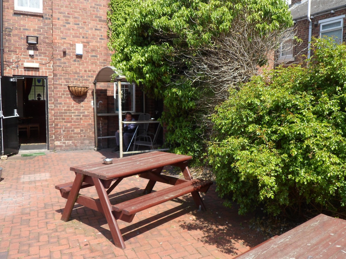 An outdoor area features a wooden picnic table surrounded by textured red brick paving. Lush green foliage provides natural cover, enhancing the inviting atmosphere of the space. The entrance to the bar is visible in the background, blending seamlessly with the surroundings.