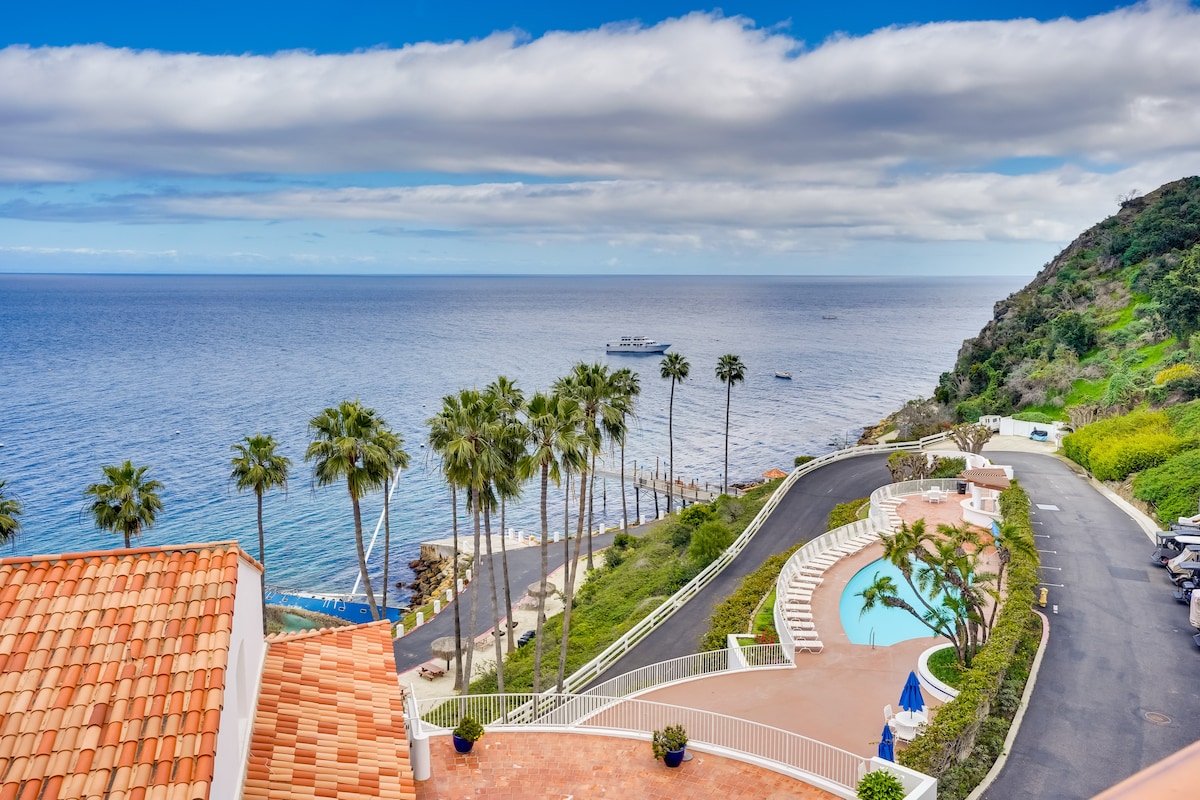 A panoramic view showcases the ocean meeting the horizon, framed by palm trees along the shore. A gently winding road leads to a pool surrounded by greenery, providing a serene and vibrant coastal atmosphere.