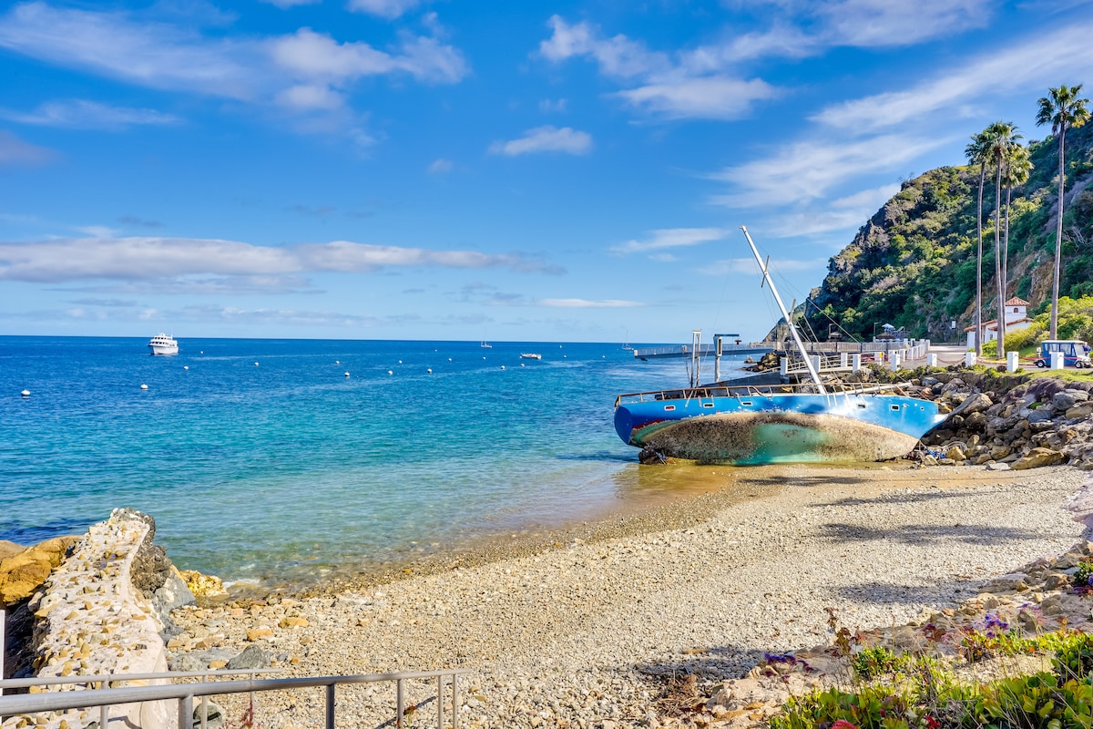 A calm beach scene is presented with clear blue waters and a rocky shoreline. A weathered boat rests on the sand, surrounded by pebbles and small rocks. Palm trees and a lush hillside frame the background, adding a natural touch to the coastal view.