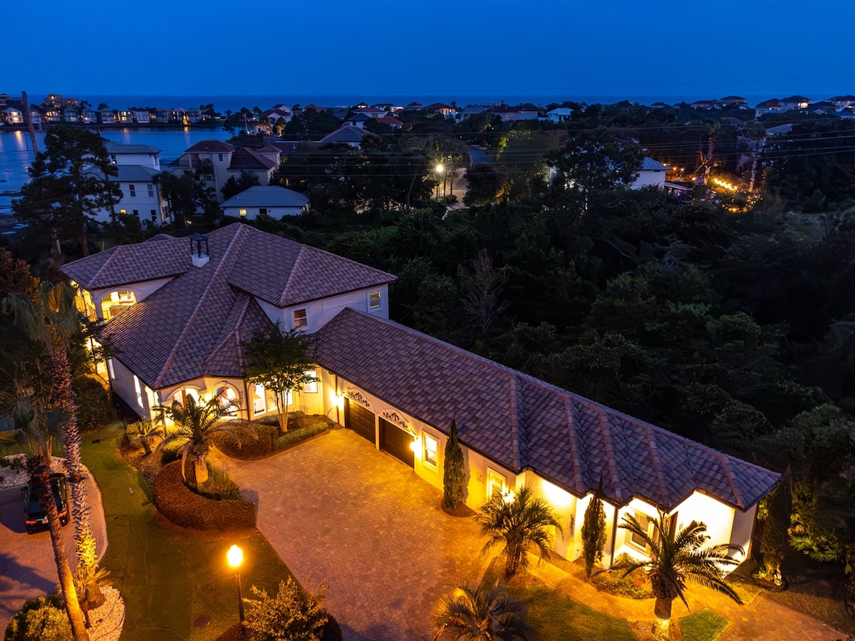 A spacious beach house is illuminated at night, showcasing a well-maintained exterior with a tiled roof and palm trees lining the entryway. The driveway leads to a garage, while distant views of water and lights from surrounding homes create a serene atmosphere.