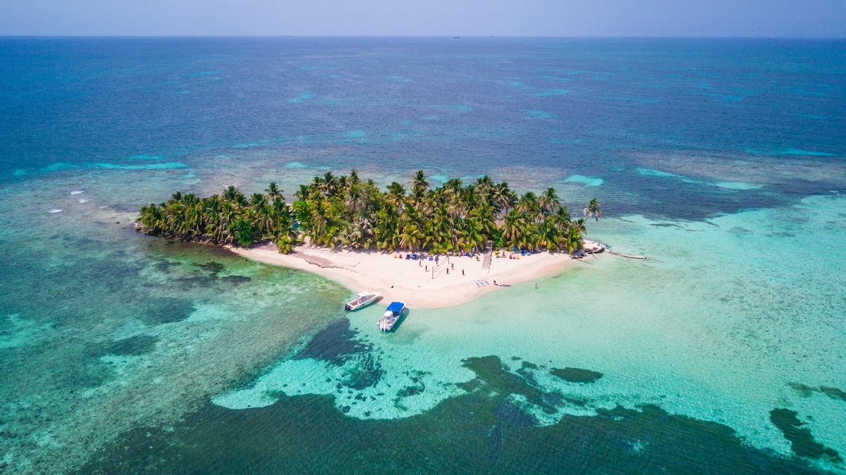 Aerial view of Ranguana Caye reveals a lush island surrounded by clear turquoise waters. Palm trees populate the sandy shore, with boats anchored nearby. The vibrant colors of the reef contrast with the white sandy beach, creating a serene tropical atmosphere.
