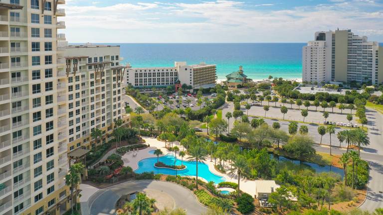A panoramic view captures the expansive lagoon-style pool surrounded by lush landscaping. In the background, the Gulf of Mexico displays its vibrant turquoise water, while pristine beaches are visible in the distance. The setting reflects the tranquility of a coastal resort.