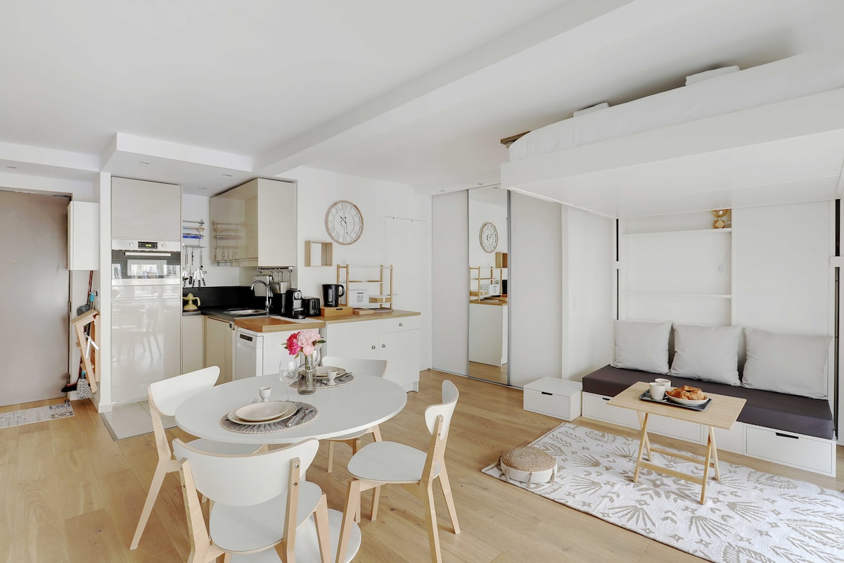 An open layout studio features a kitchen with modern appliances and a dining area with a round table and light-colored chairs. A comfortable seating area is accompanied by a coffee table, while a bed is tucked neatly above, optimizing space in the room.