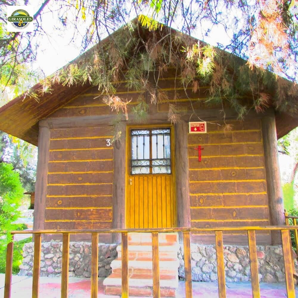 A rustic cabin with a wooden façade features a bright yellow door, framed by a decorative iron window. Natural stone accents around the foundation add texture, while a wooden railing encloses the porch area, providing a welcoming entrance.
