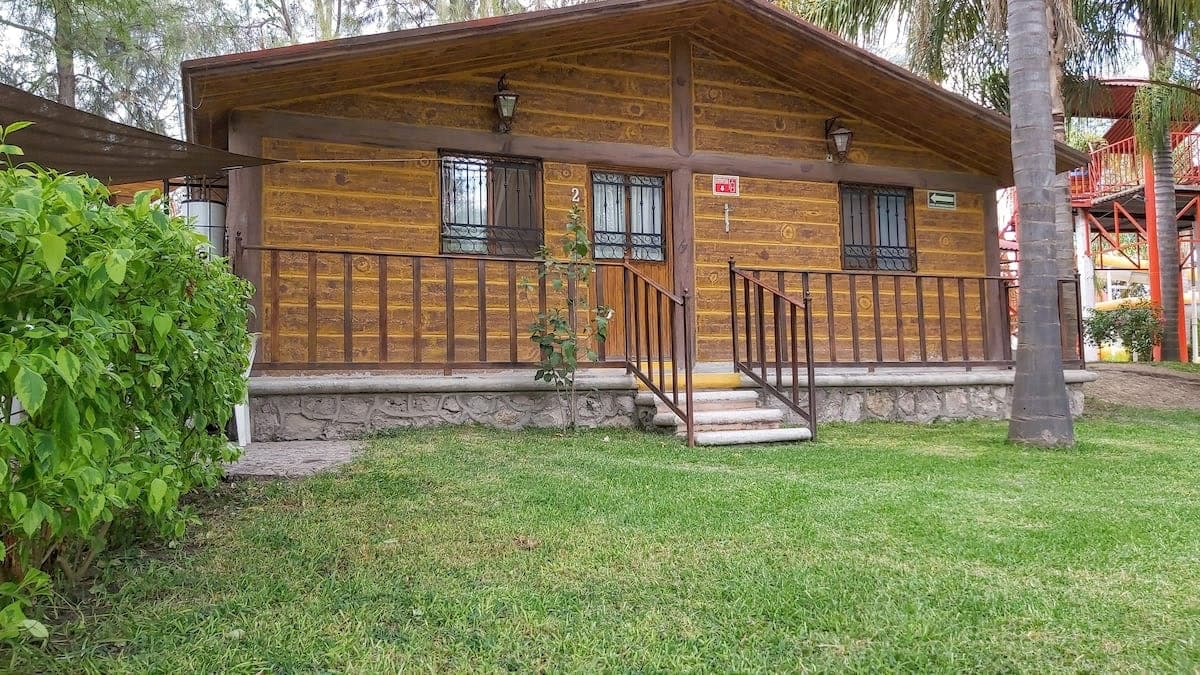 A charming wooden cabin features a welcoming entryway with a stone pathway leading to the front steps. The exterior is framed by lush green grass and shrubs, with decorative lighting highlighting the cabin’s rustic design.