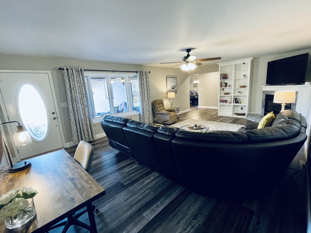 A cozy living room features a large sectional sofa facing a modern television and decorative shelves. Natural light filters through large windows adorned with patterned curtains. A dining table is positioned nearby, offering a space for meals or gatherings.