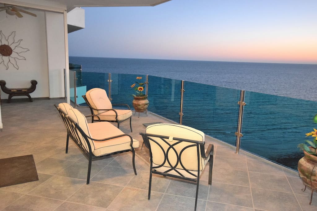 A spacious balcony overlooks the ocean, featuring two comfortable lounge chairs and a small table. The glass railing provides unobstructed views of the calm sea, with hues of blue reflecting the evening sky. Planters with vibrant flowers enhance the outdoor space.