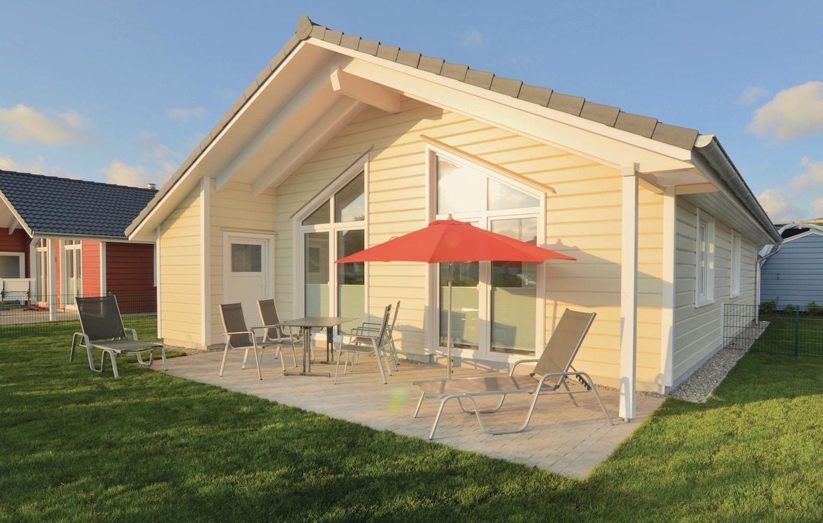 A detached holiday home is presented with large glass windows allowing natural light to fill the interior. The terrace features an umbrella over a table surrounded by several loungers, offering a space for relaxation. The well-maintained grass adds to the outdoor comfort.
