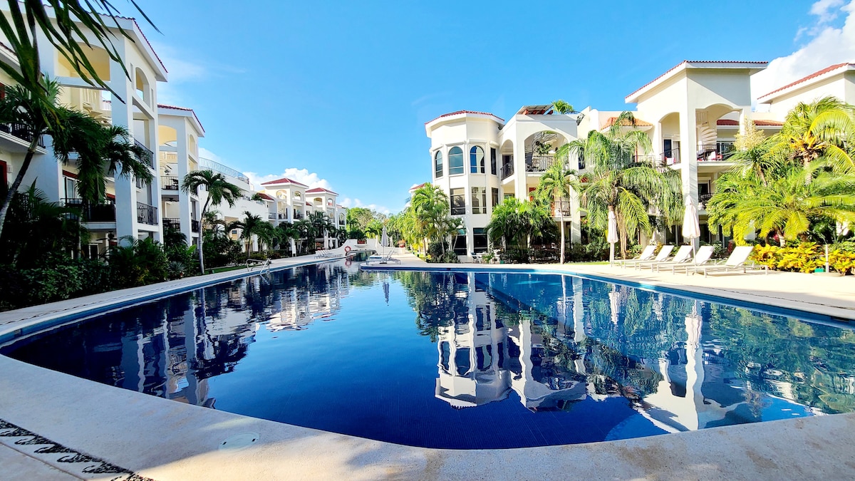 A spacious outdoor swimming pool reflects the surrounding palm trees and blue sky. Modern buildings frame the pool area, with lounge chairs positioned along the edge. Lush greenery adds a touch of nature, enhancing the tranquil setting.