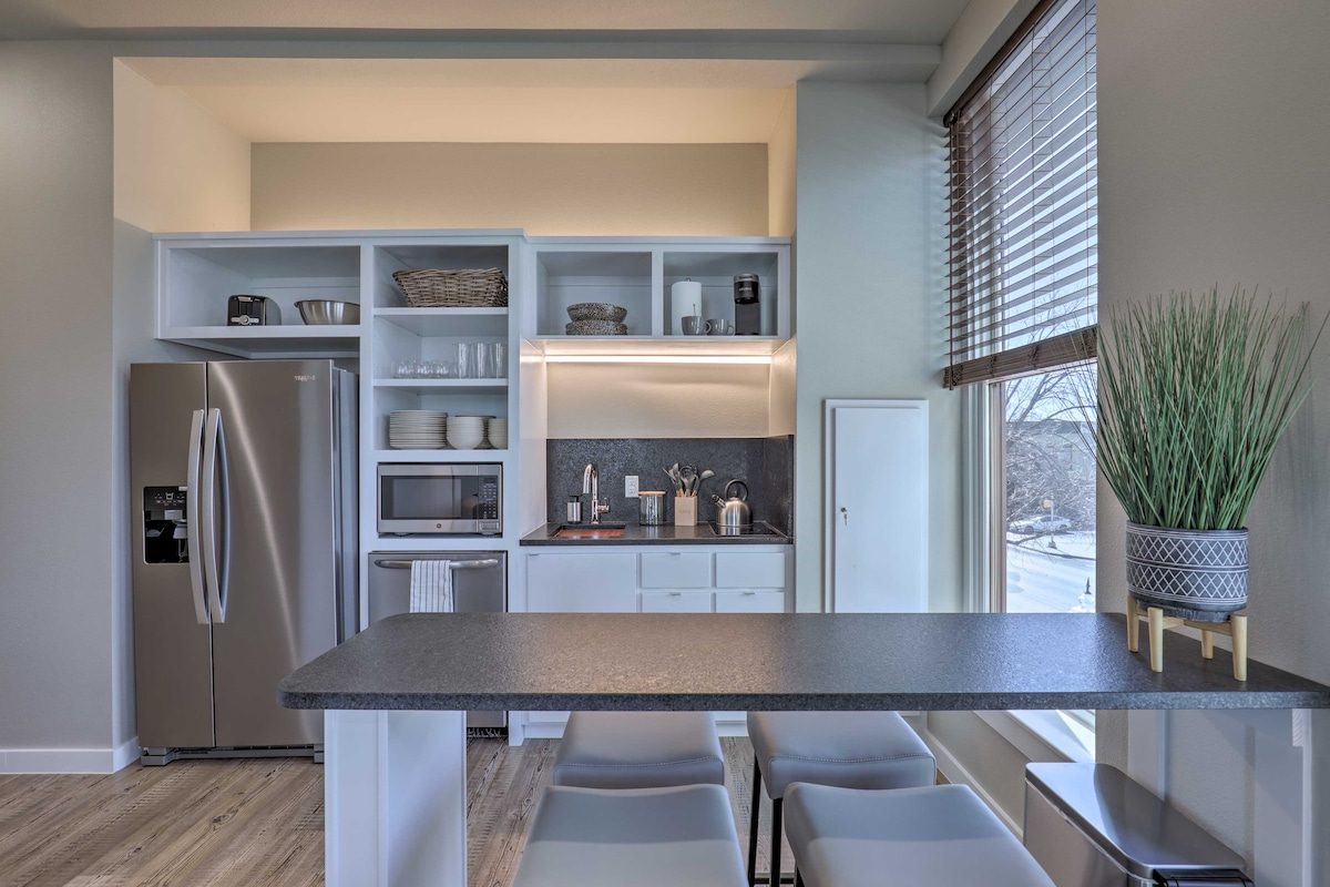 A modern kitchenette is displayed with sleek cabinetry, stainless steel appliances, and a countertop with seating. A window offers natural light, while open shelves showcase dishware and utensils. A small potted plant adds a touch of greenery to the space.