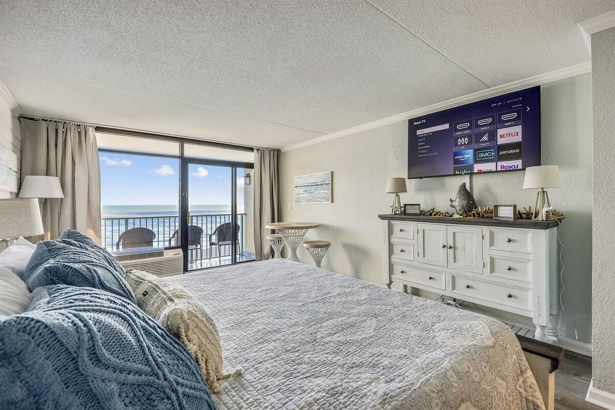 A serene bedroom space features a spacious bed with soft linens and decorative pillows. A large flat-screen TV mounted on the wall is complemented by a stylish dresser. Floor-to-ceiling windows provide views of the ocean and access to the private balcony.