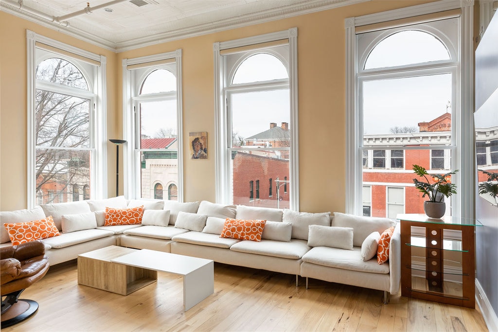 A spacious living area features large, arched windows that allow natural light to fill the room. A wrap-around couch adorned with orange accent pillows invites relaxation, while a light wooden coffee table sits in the center. The hardwood flooring enhances the open and airy ambiance.