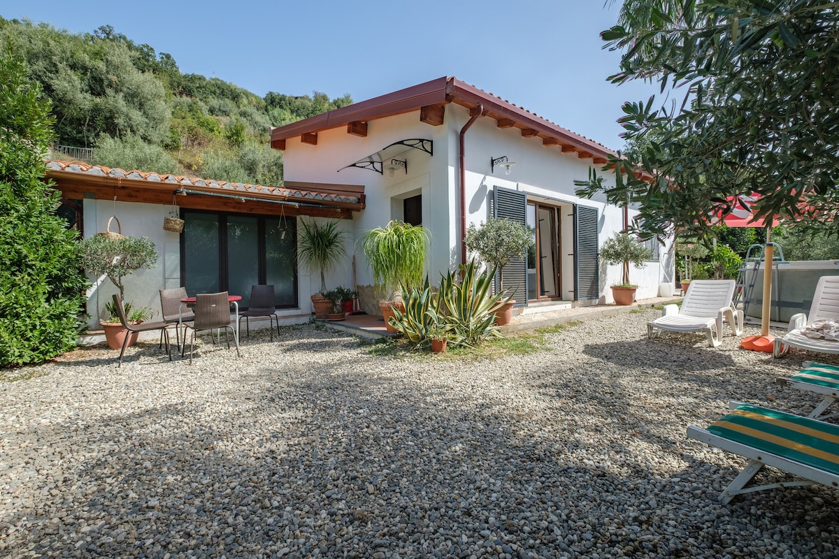 The exterior of the Casa di Martina is presented, featuring a gravel patio surrounded by greenery. The white building has large windows and a covered entrance. Sun loungers and potted plants are visible, adding to the inviting outdoor space.
