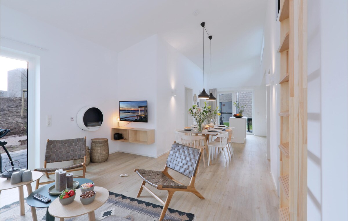 A modern living area features two wooden lounge chairs with woven seats, accompanied by a small coffee table. A flat-screen television is mounted on the wall, and a dining area with a light wood table is visible in the background, illuminated by natural light from large windows.