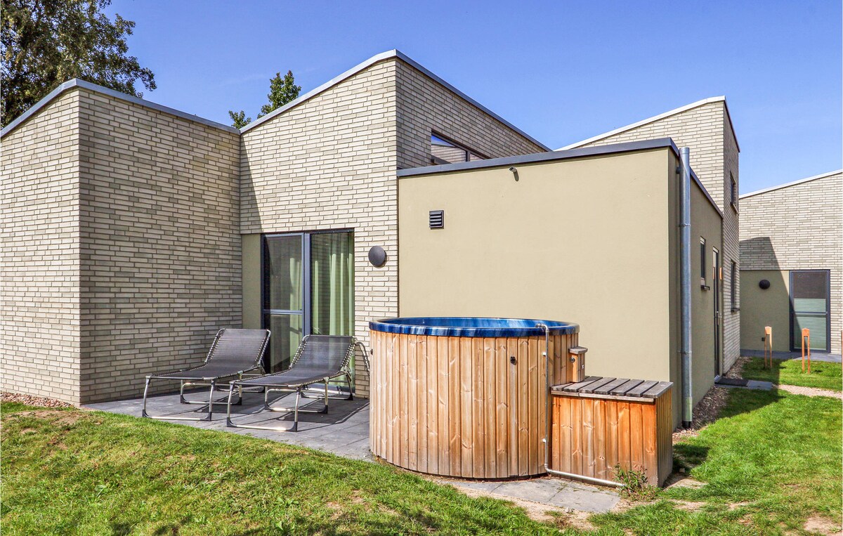 An outdoor area features a wooden hot tub on a stone patio. Two lounge chairs are positioned nearby, with a grassy area surrounding the space. Natural light brightens the setting, highlighting the modern architectural design of the building in the background.