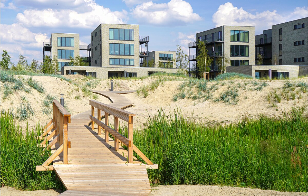 A wooden bridge leads over lush green grass and sandy terrain toward modern multi-story buildings. The structures feature large windows and balconies, surrounded by greenery that enhances the sense of natural beauty in the area.