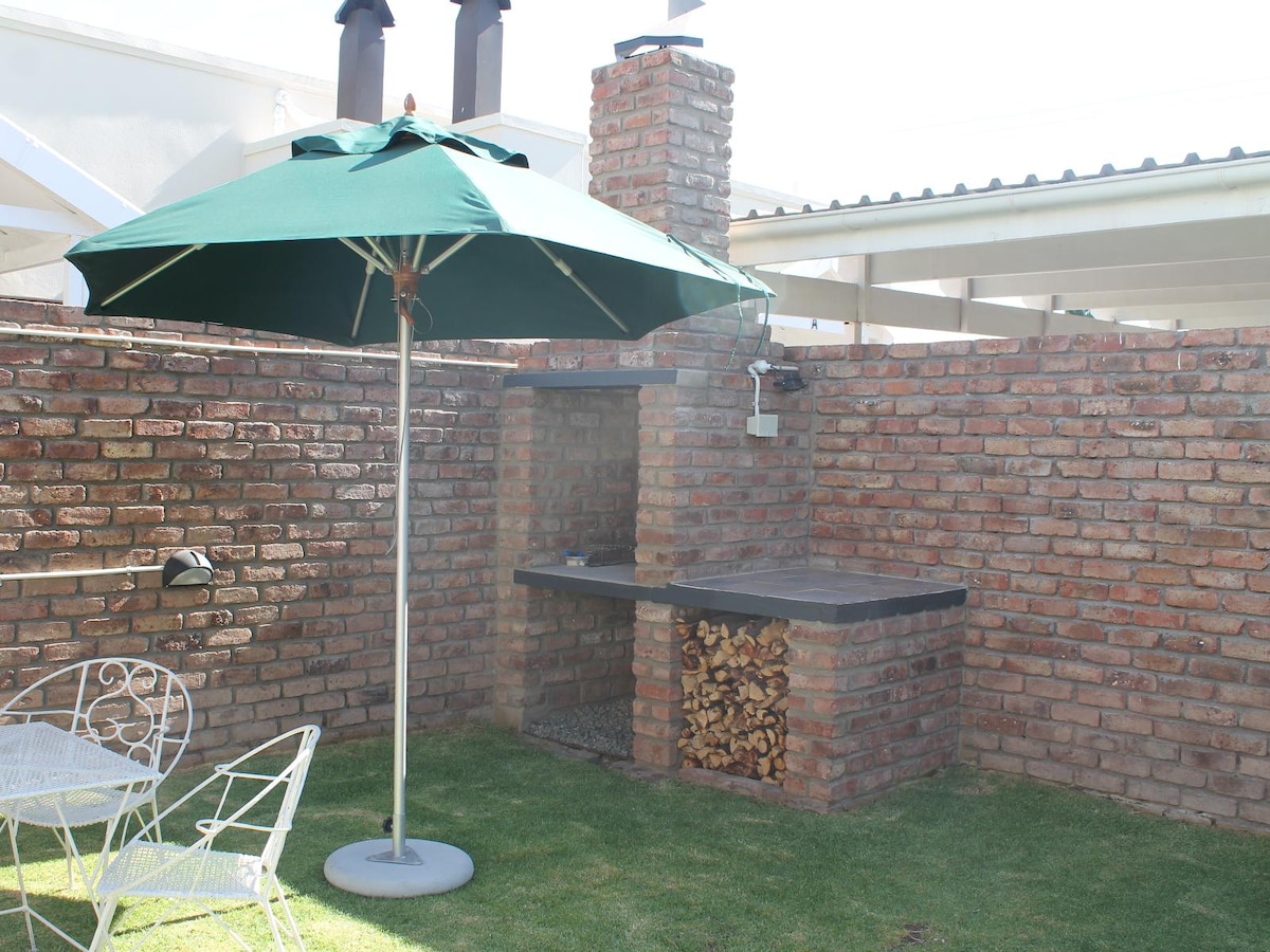 An outdoor braai area is surrounded by a sturdy brick wall, featuring a built-in barbecue and a small stack of firewood. A large green umbrella provides shade over a round table with two white chairs, situated on a well-kept lawn.