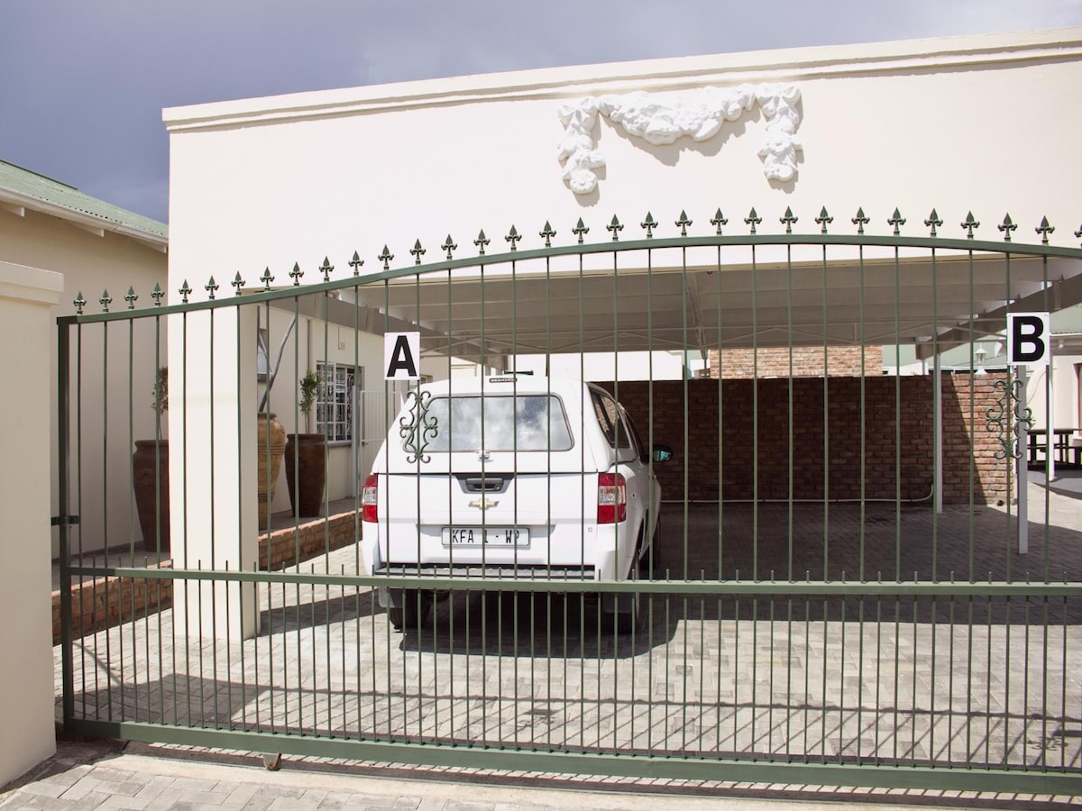 A gated parking area is visible, providing undercover space for vehicles. The entrance features a decorative green metal gate with pointed tops. A light-colored vehicle is parked inside, with brick walls and planters adding structure to the surroundings.