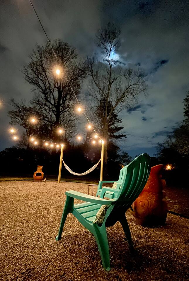 An outdoor space is available, featuring a turquoise chair under a canopy of lights strung between trees. A hammock is suspended nearby, creating a cozy leisure area. The gravel ground and distant fire pit enhance the inviting atmosphere, with a partially cloudy night sky above.