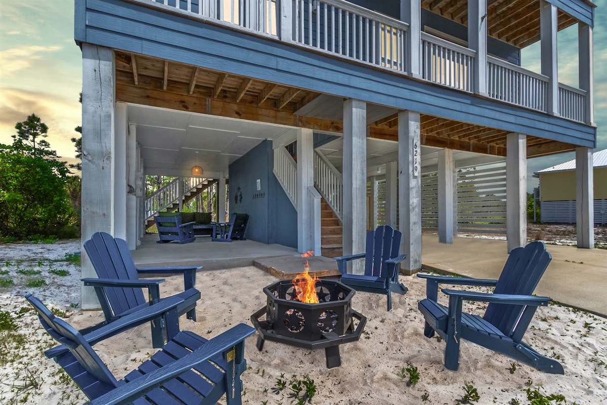 A cozy fire pit area is surrounded by blue adirondack chairs, set on a sandy ground. The elevated home above provides a welcoming outdoor space with access to seating under the structure. Soft lighting indicates an inviting atmosphere for evening gatherings.