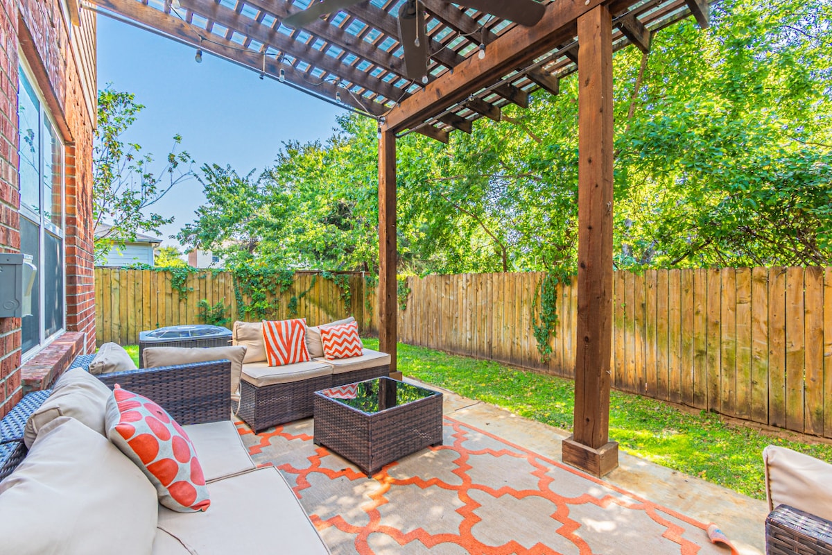 A shaded outdoor patio features comfortable seating with orange and cream accent pillows. A glass-top coffee table is centered on a patterned rug, surrounded by lush greenery and a wooden fence. Sunlight streams gently through the overhead structure, creating a welcoming relaxation space.