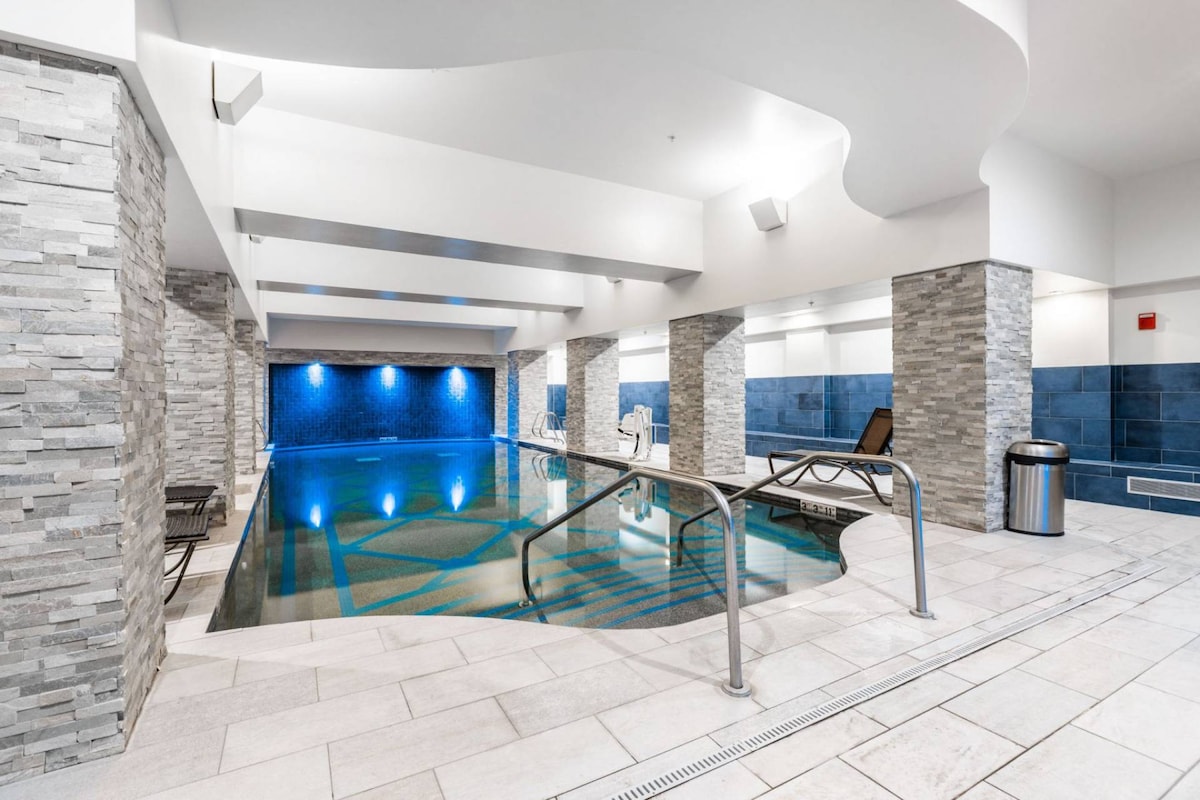 An indoor pool area is highlighted by modern architectural features, including elegant stone walls and soothing blue lighting. Lounge chairs are arranged around the pool, which is designed with a gentle gradient and tiled patterns, creating a relaxing ambiance.