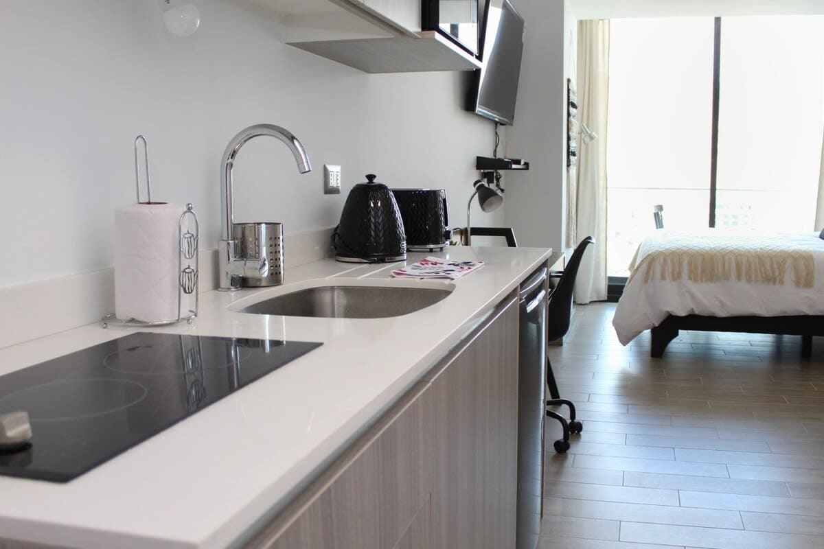 A modern and fully equipped kitchen is displayed, featuring a sleek countertop, sink, and stovetop. A coffee maker and kettle are positioned nearby, while a smart TV is mounted above. A stylish dining chair is visible next to the workspace, with ample natural light filling the area.