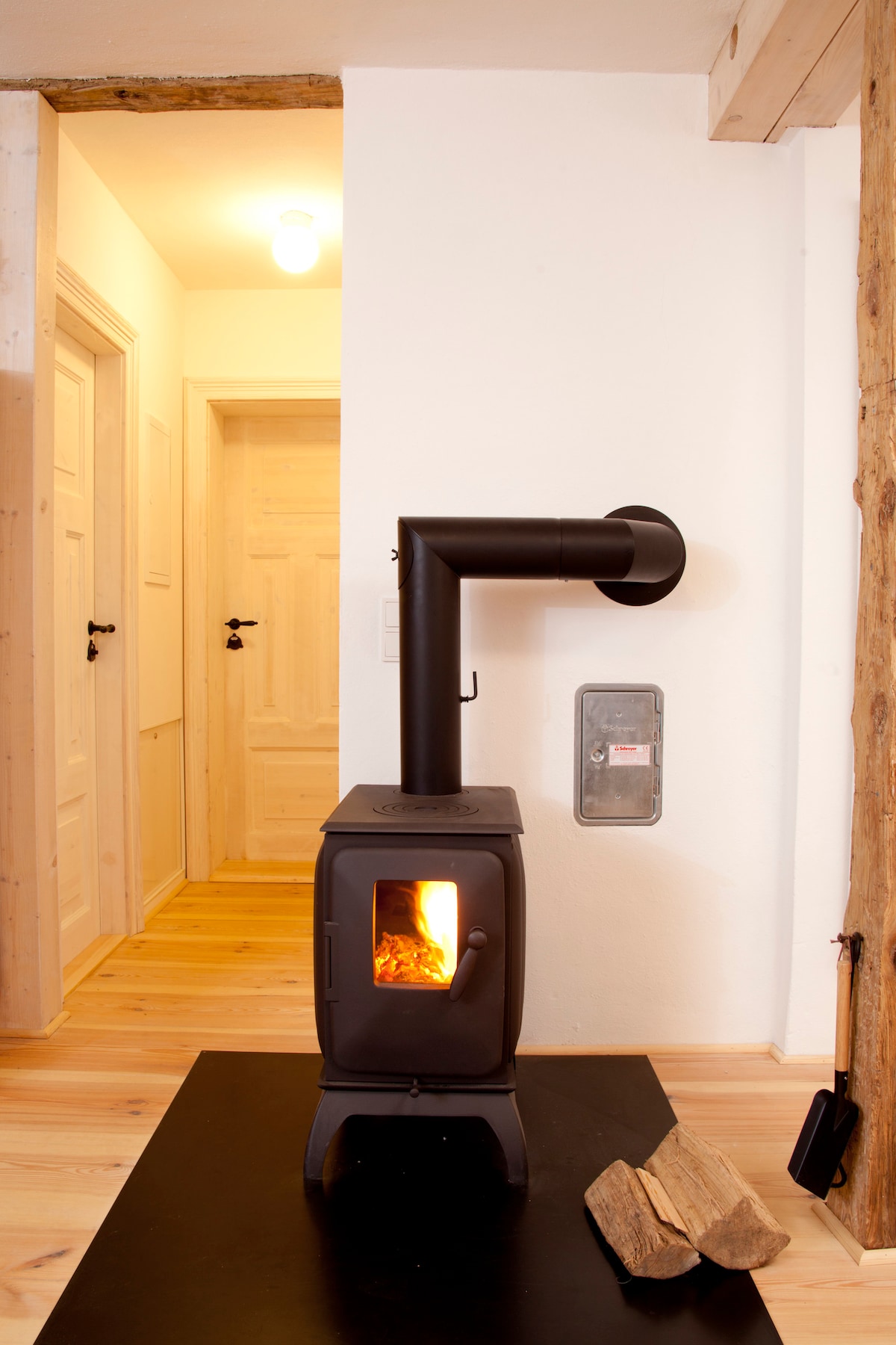 A compact wood stove is situated against a white wall, providing warmth with a visible flame. Light wood flooring and a doorway can be seen in the background, leading to a well-lit hallway with wooden doors.