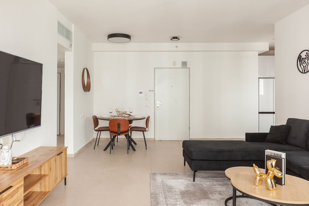 A spacious living area is highlighted by a large black sectional sofa opposite a wall-mounted television. A round dining table for four is positioned nearby, with minimalist decor enhancing the modern aesthetic. Natural light fills the room through expansive windows, complementing the sleek, neutral color palette.