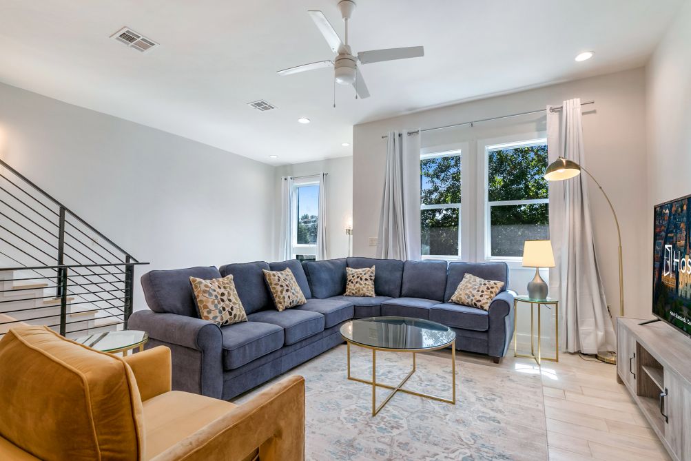 The living room features a spacious sectional sofa with decorative pillows, complemented by a glass coffee table. A ceiling fan and modern lamp provide overhead lighting. Large windows fill the area with natural light, highlighting neutral walls and hardwood floors.