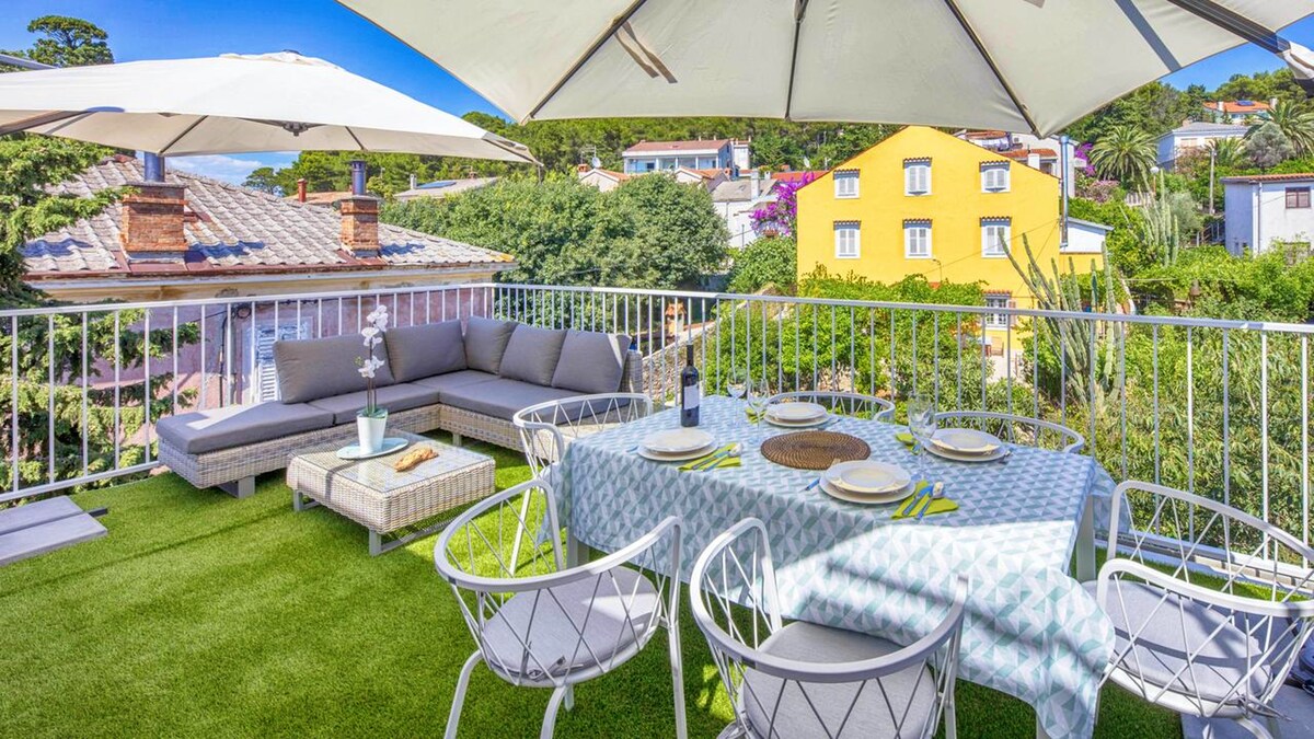 An outdoor terrace is displayed with a shaded seating area featuring a large sofa and a dining table set for meals. Umbrellas provide shade over the space, surrounded by lush greenery and colorful buildings in the distance, creating a relaxing setting.