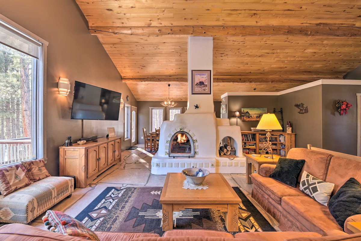 The spacious living room features a double fireplace built into a white adobe wall. Comfortable seating, including a large sofa, is arranged around a wooden coffee table. Natural light floods the space through large windows, illuminating the vaulted wood ceiling.
