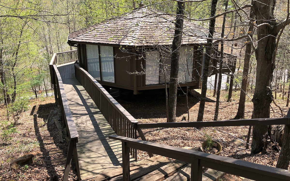 A treehouse-style chalet is nestled among lush greenery, featuring a circular shape with large windows that bring in natural light. A wooden ramp leads to the entrance, flanked by sturdy rails, inviting access to the spacious outdoor deck that wraps around the home.