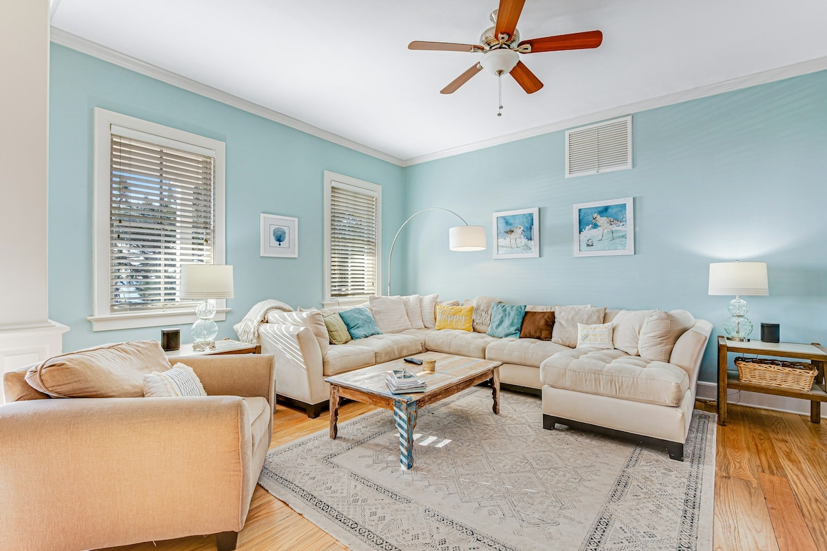 A spacious living area features a large, L-shaped sectional sofa adorned with assorted cushions in various colors. Light wood flooring complements the soft blue walls, while natural light filters through the window blinds. Two side tables with lamps provide additional lighting, enhancing the room's inviting layout.