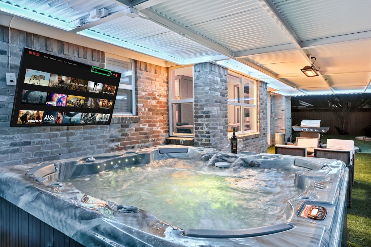 A private hot tub is situated under a covered patio, featuring soft lighting and a sleek design. A mounted TV is visible, displaying streaming options. The surrounding brick walls and outdoor seating create a cozy atmosphere for relaxation and entertainment.
