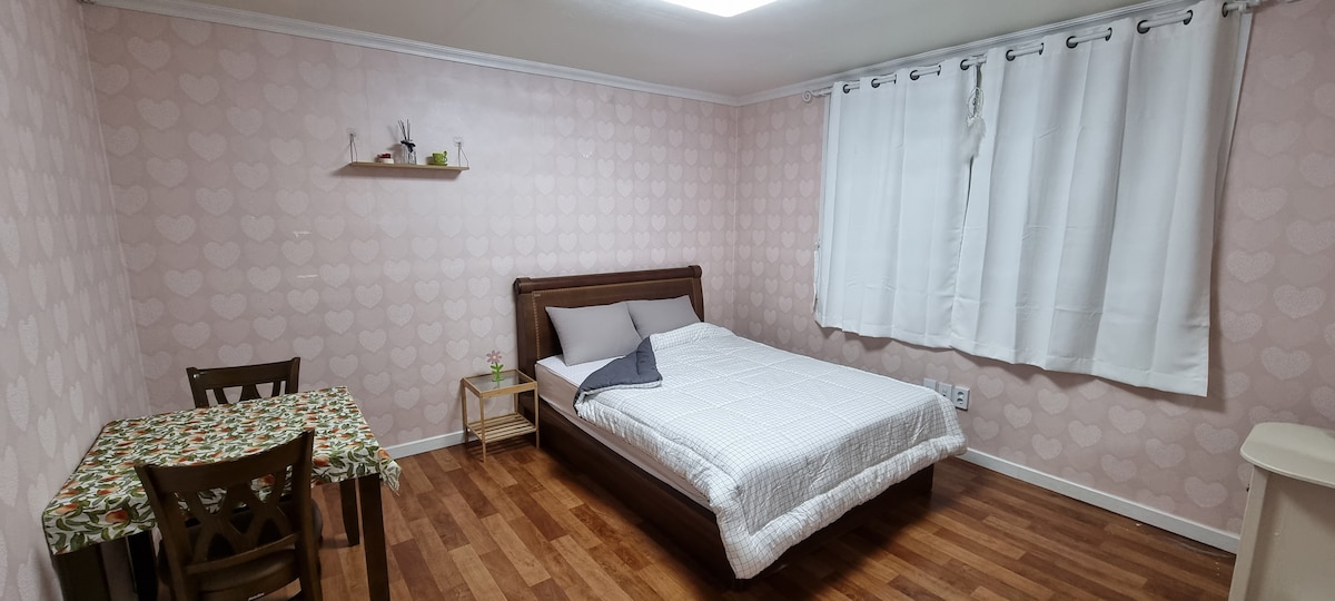A cozy sleeping area features a spacious bed with neutral bedding set against a subtly patterned pink wall. A small wooden table and chairs are positioned nearby, complemented by a light-colored window treatment allowing natural light to brighten the room.