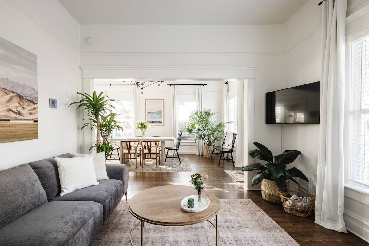 A bright living area showcases a spacious gray couch positioned next to a round wooden coffee table. A large wall-mounted TV is visible, while a dining space with plants and a wooden table is seen through an adjacent room. Natural light fills the space.
