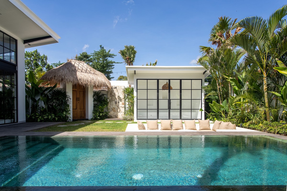 A spacious outdoor area features a rectangular pool with clear water, surrounded by tropical greenery. A thatched-roof hut is positioned adjacent to a modern structure with large glass windows. Cushions are neatly arranged along the pool edge, enhancing the relaxing atmosphere.
