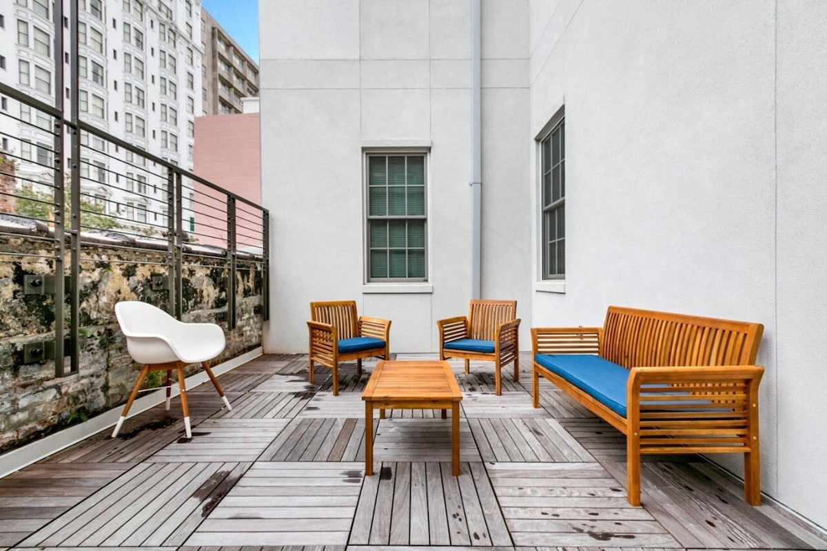A spacious patio area features wooden furniture including two armchairs, a loveseat, and a coffee table. A white mid-century modern chair is positioned in the corner. The wooden deck is surrounded by building walls, with large windows visible in the background.