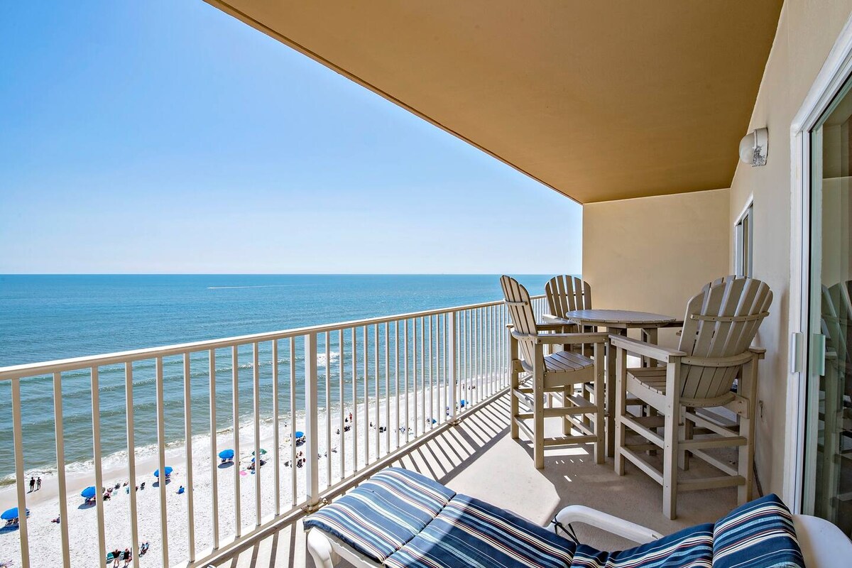 A spacious private balcony offers sweeping views of the Gulf, featuring a table surrounded by high-backed chairs. Comfortable lounge chairs are positioned for relaxation, and the sandy beach can be spotted below alongside colorful beach umbrellas.