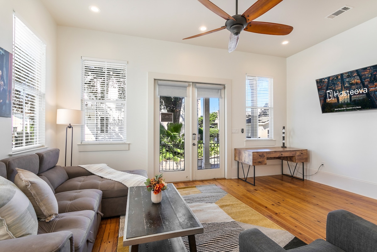 A living area features a sectional sofa with a throw blanket draped neatly. Natural light floods through multiple windows, illuminating the wooden flooring. A decorative coffee table holds a small potted plant. Two set of glass doors provide access to an outdoor space, while a wall-mounted TV is present.
