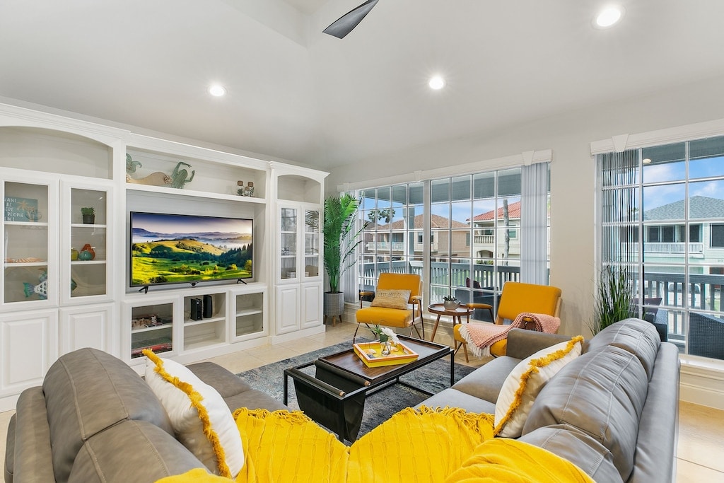 A spacious living room is adorned with a large sectional sofa and accent chairs, featuring a wall-mounted TV and natural light from expansive windows. Decorative shelves hold various items, complemented by indoor plants, creating a cozy yet modern ambiance.