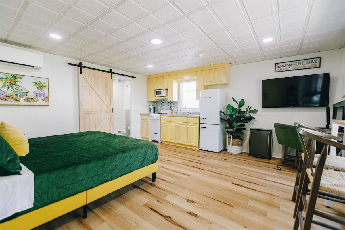 The studio features a king-sized bed with a green coverlet and vibrant yellow accents. A full kitchen is visible with bright yellow cabinets and modern appliances. A flat-screen TV is mounted on the wall, and natural wood floors enhance the spacious layout.