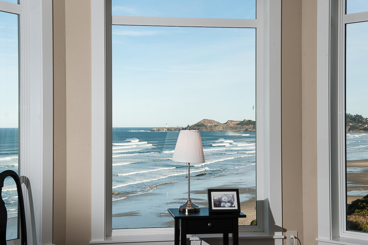 Large windows frame a stunning view of the Pacific Ocean and shoreline, showcasing rolling waves and coastal hills. A small table with a lamp and a picture frame anchors the space, enhancing the serene atmosphere projected by the scenic vista.